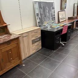 Tweedehands meubels uitgestald: houten kasten, zwarte bureaus en een roze stoel in een kringloopwinkel