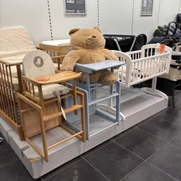 Uitgestalde babymeubels en accessoires: witte en houten babybedje, kinderstoel, grote teddybeer en verschillende baby-artikel
