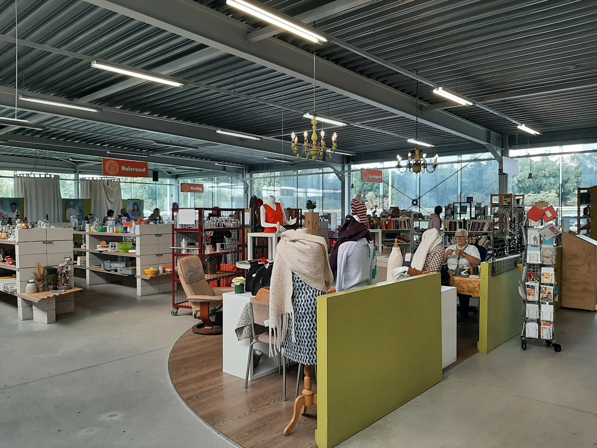 Winkelvloer van kringloopwinkel met groene scheidingswanden, boekenrekken, stellingen en bezoekers die winkelen