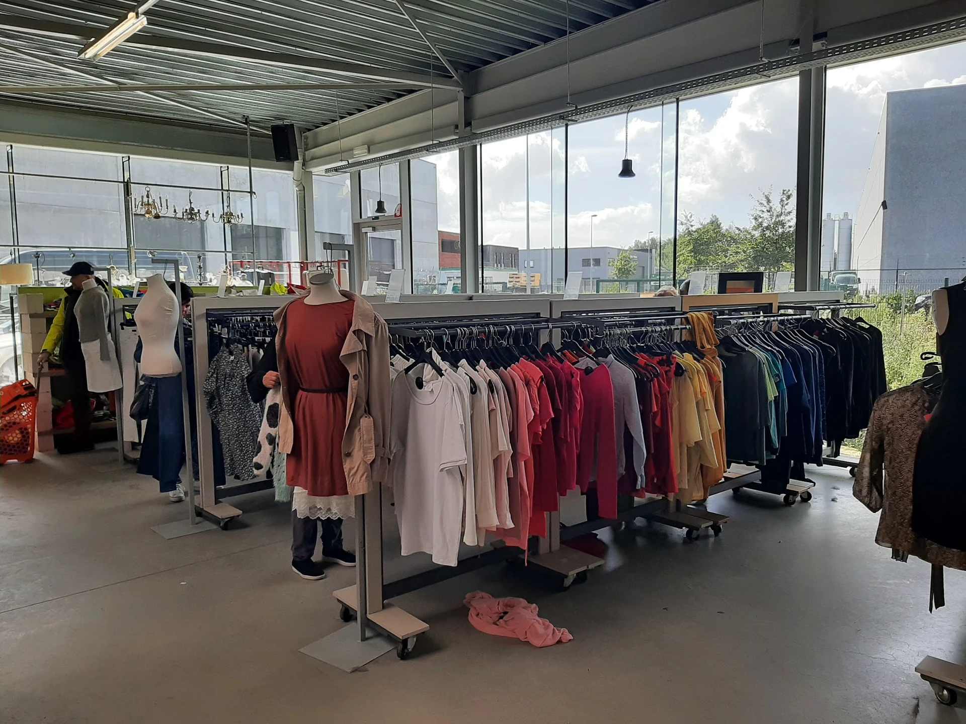Rekken met tweedehands kleding in verschillende kleuren en stijlen in een winkelruimte met grote ramen.