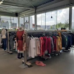 Rekken met tweedehands kleding in verschillende kleuren en stijlen in een winkelruimte met grote ramen.