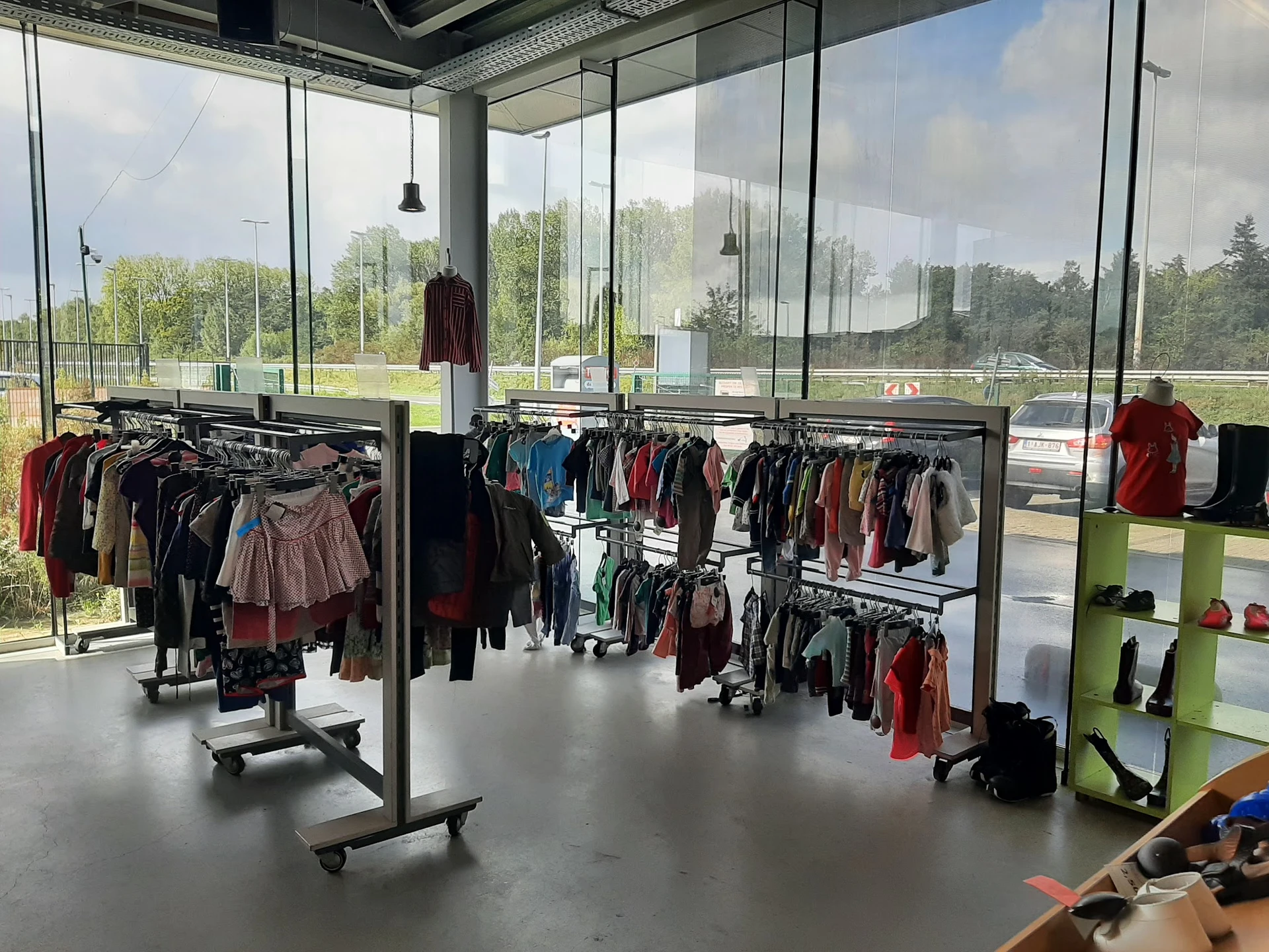 Winkelinterieur met kledingrekken volgeladen met tweedehands kleding, grote glazen ramen en uitzicht op groen terrein