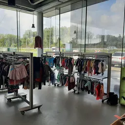 Winkelinterieur met kledingrekken volgeladen met tweedehands kleding, grote glazen ramen en uitzicht op groen terrein