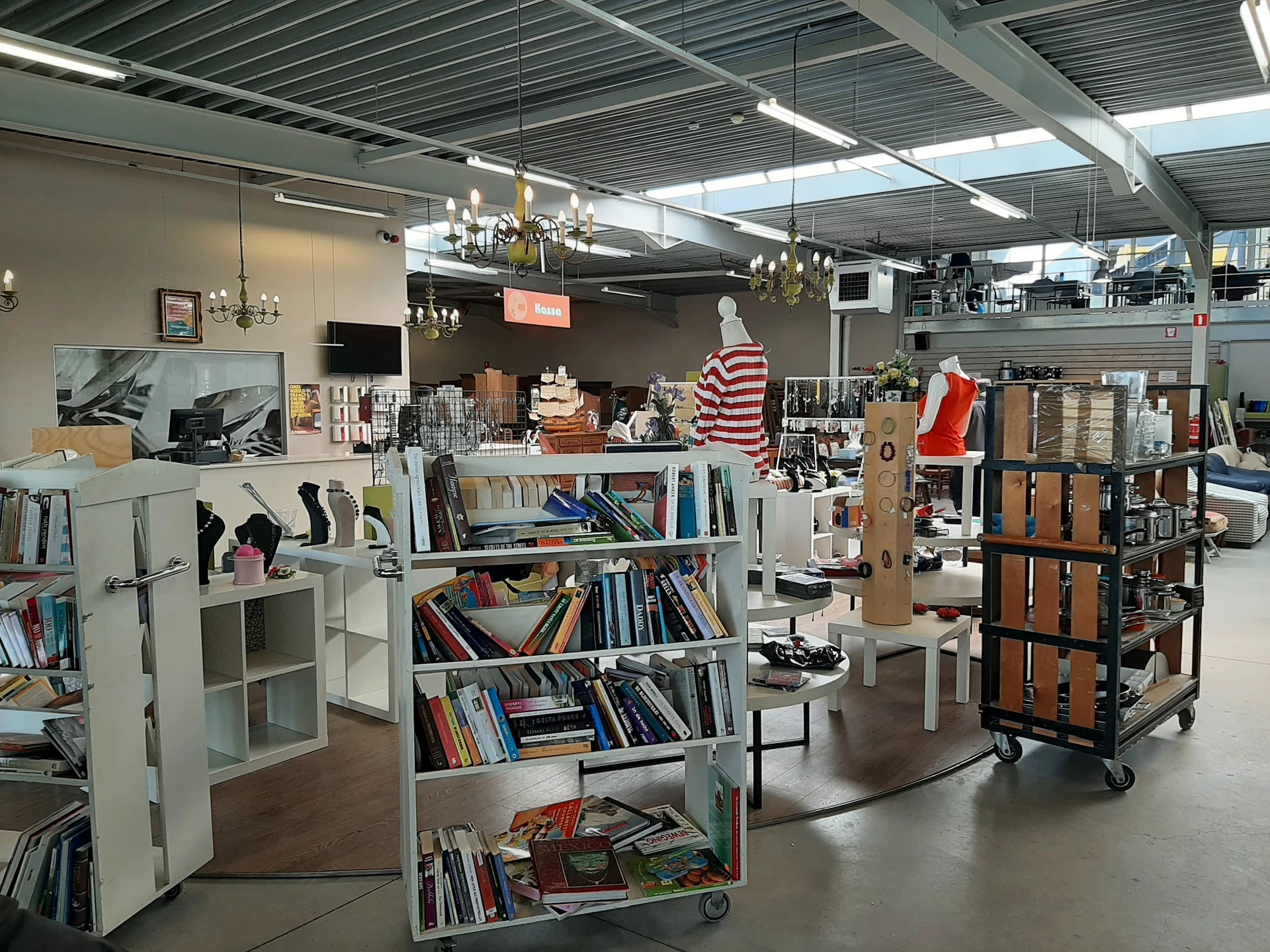 Winkelruimte met boekenkasten, displaykasten, hangende decoraties en diverse tweedehands artikelen onder industrieel plafond