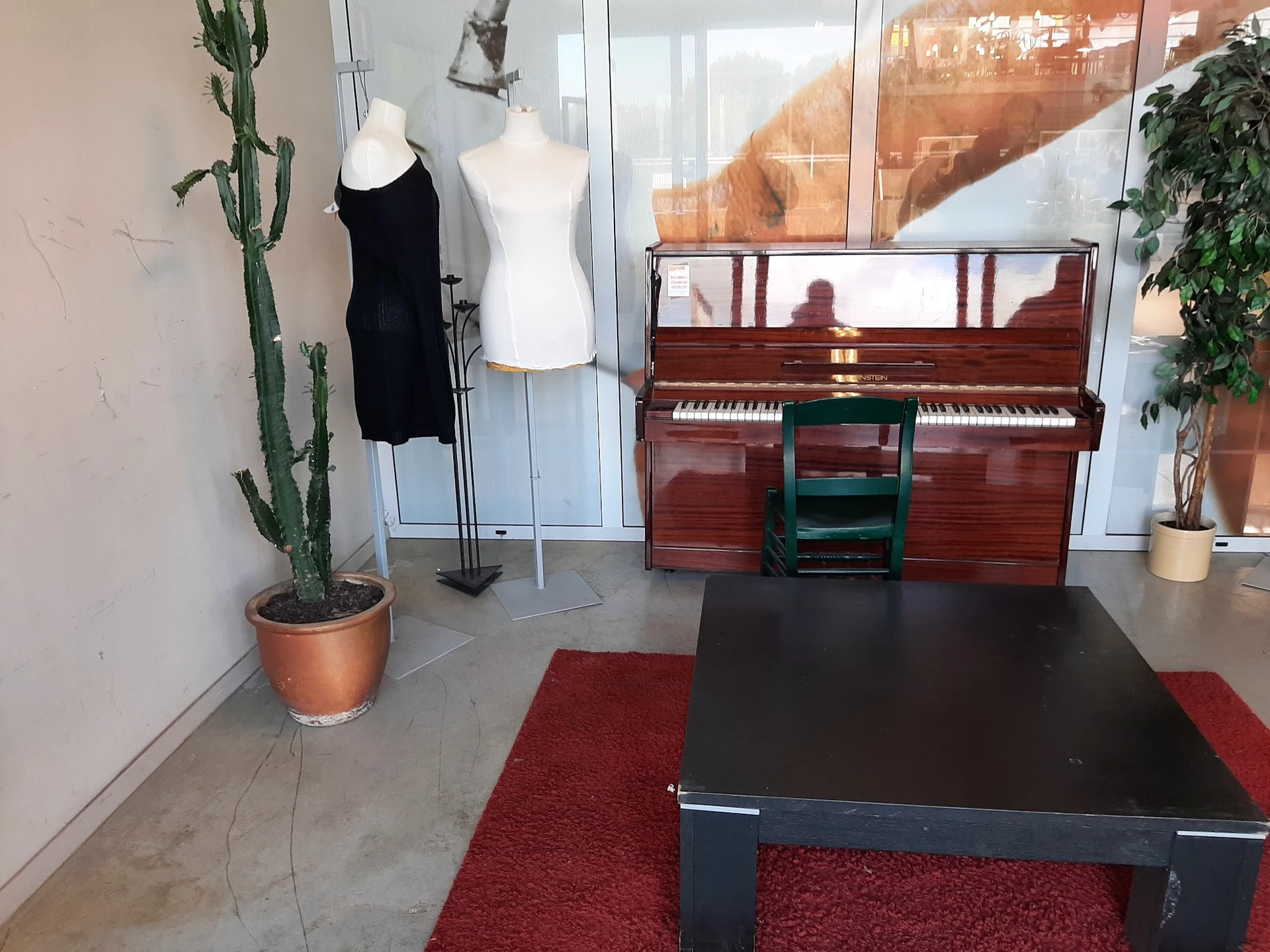 Winkelinterieur met rode piano, kledingpoppen, cactus en rode loper op betonnen vloer