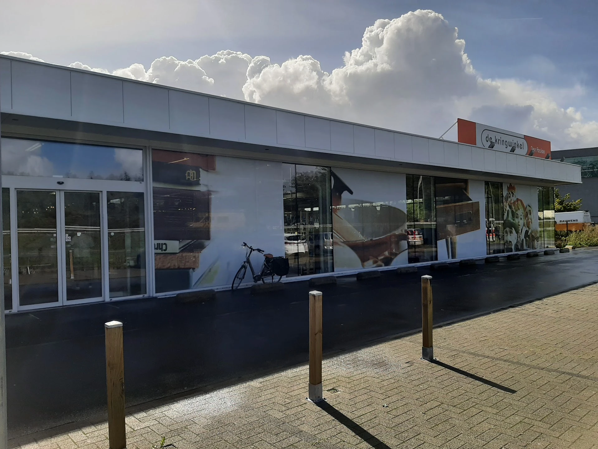 Witte gevel van kringloopwinkel met grote ramen, zwarte sokkel en houten paaltjes op het terras