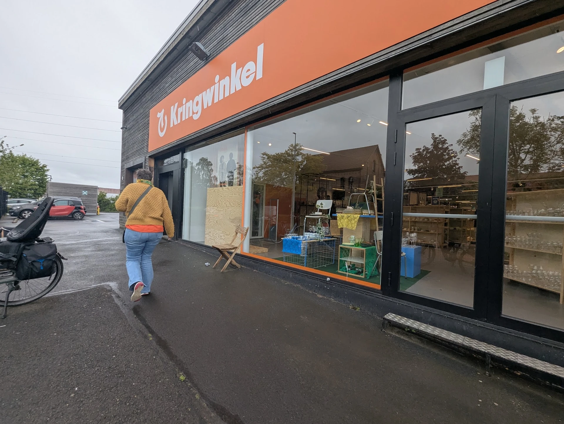 Gevel van kringloopwinkel met oranje/rode pui en grote ramen, winkelname leesbaar, persoon in gele jas loopt voorbij