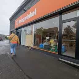 Gevel van kringloopwinkel met oranje/rode pui en grote ramen, winkelname leesbaar, persoon in gele jas loopt voorbij