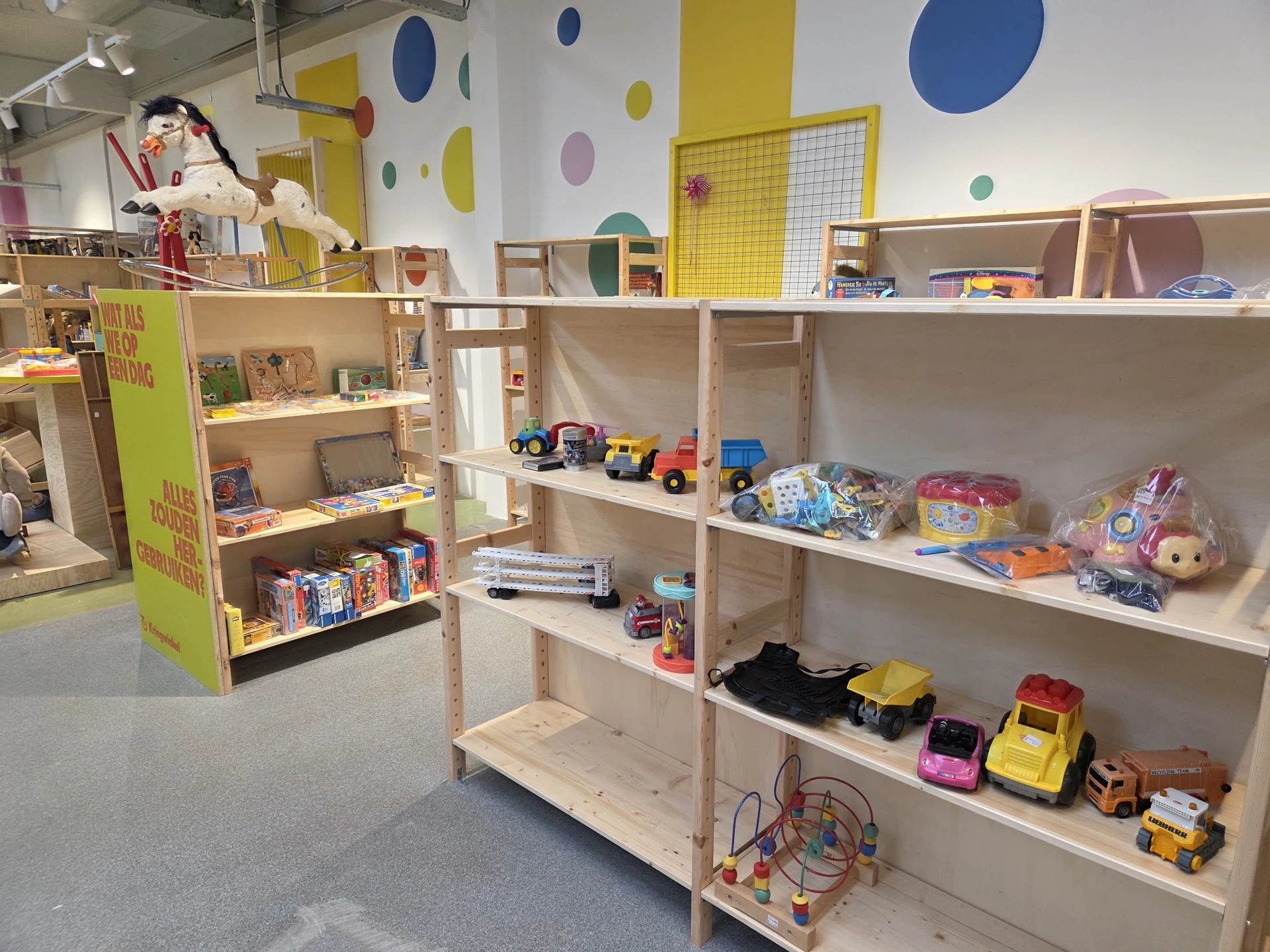 Speelgoed en kinderspullen op houten stellingen in een kringloopwinkel, met gekleurde cirkels aan het plafond