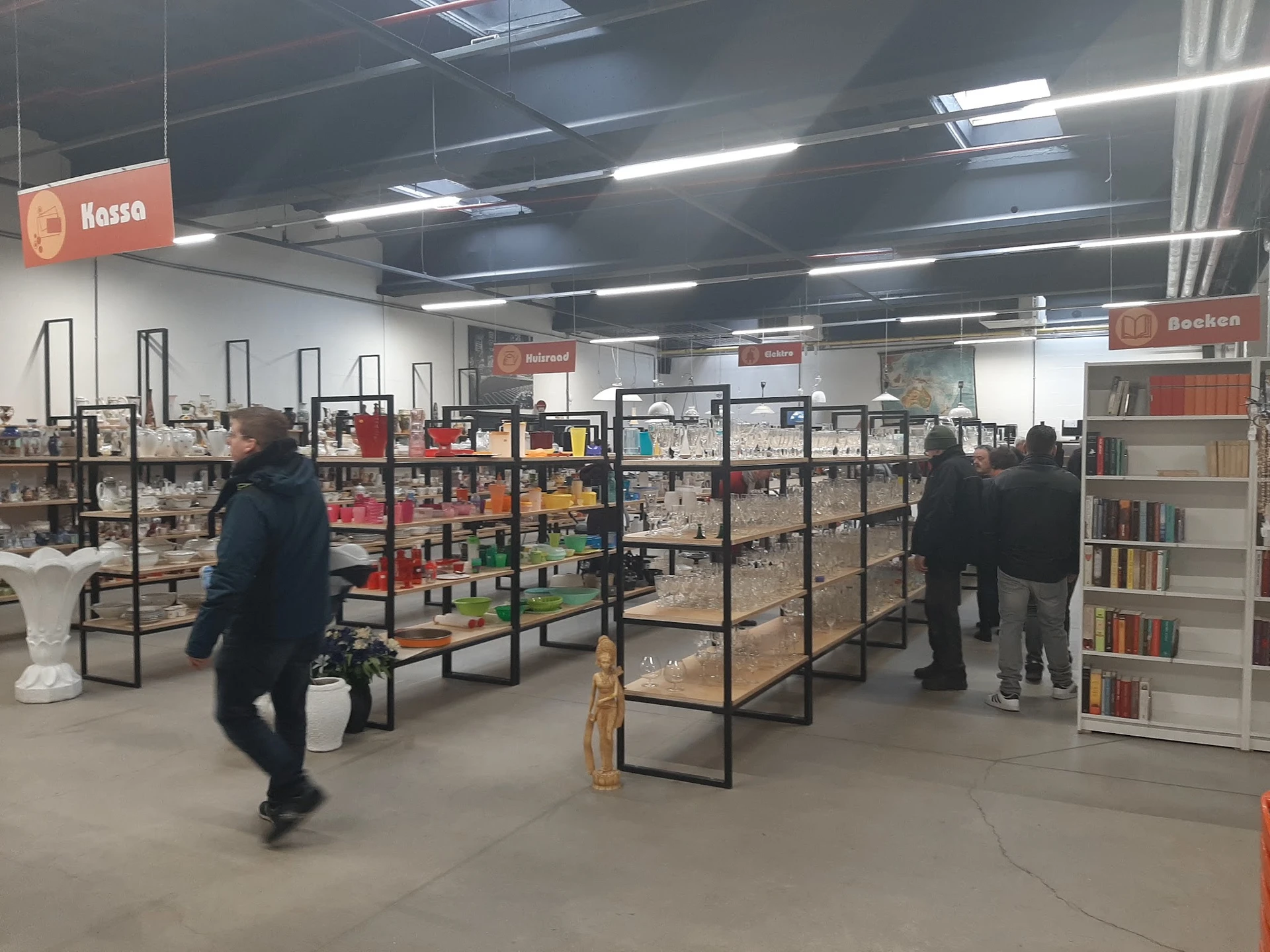 Winkelvloer met metalen stellingen gevuld met tweedehands artikelen, klanten winkelen, tl-verlichting aan plafond