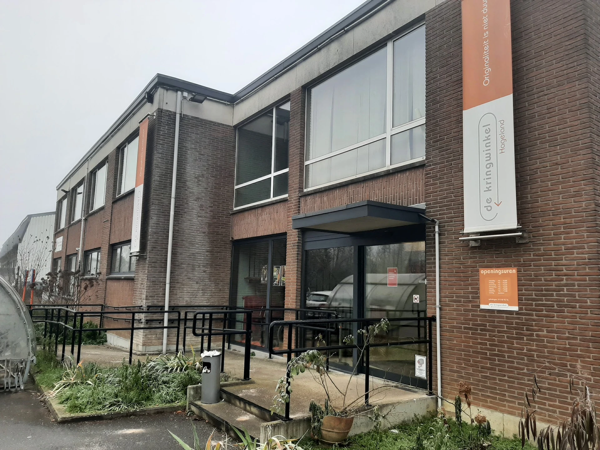 Baksteinen gevel van kringloopwinkel met grote ramen, ingang met zwarte trapleuning en oranje decoratieve elementen