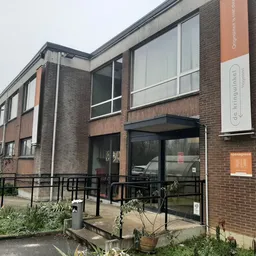 Baksteinen gevel van kringloopwinkel met grote ramen, ingang met zwarte trapleuning en oranje decoratieve elementen