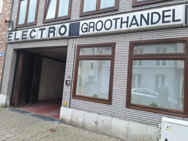 Gevel van kringloopwinkel met 'ELECTRO GROOTHANDEL' uithangbord, bruine houten ramen en open deur