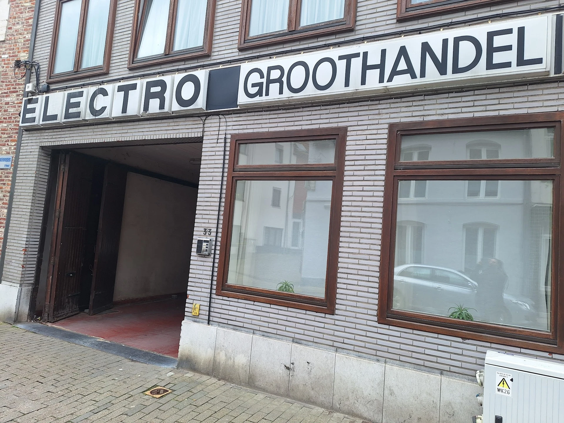 Gevel van kringloopwinkel met 'ELECTRO GROOTHANDEL' uithangbord, bruine houten ramen en open deur