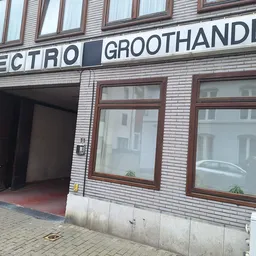 Gevel van kringloopwinkel met 'ELECTRO GROOTHANDEL' uithangbord, bruine houten ramen en open deur