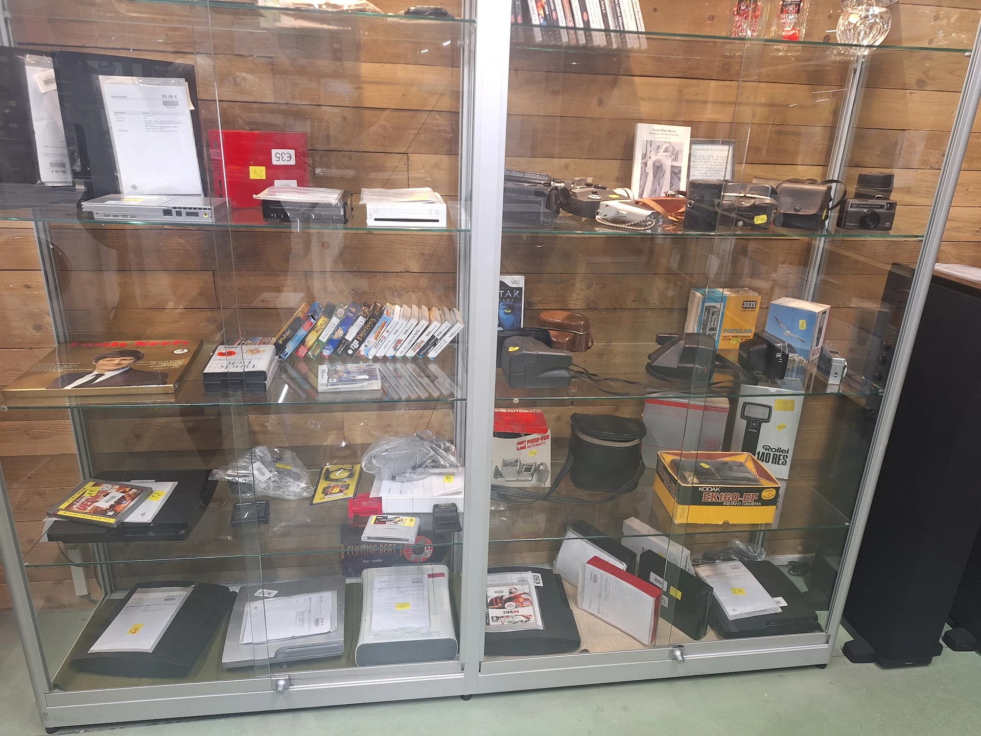 Glazen vitrinekast met diverse tweedehands elektronische apparaten, boeken, gamingartikel en andere items