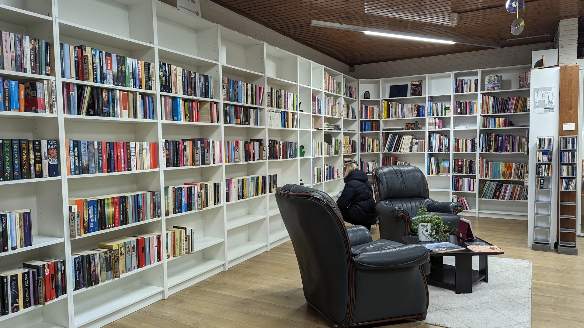Boekenrek met grote witte kasten vol boeken, zwarte leunstoel en plant in kringloopwinkel