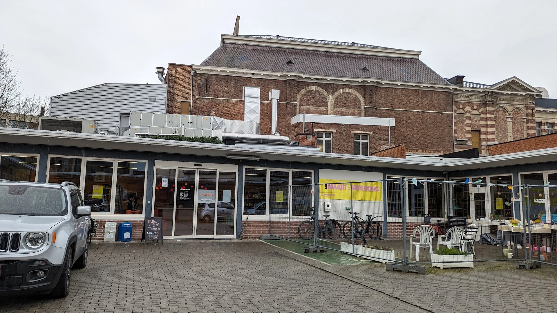 Voorgevel van kringloopwinkel met bakstenen pand en moderne winkelpui met ramen, fiets tegen zijkant, terras met stoelen