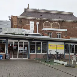 Voorgevel van kringloopwinkel met bakstenen pand en moderne winkelpui met ramen, fiets tegen zijkant, terras met stoelen