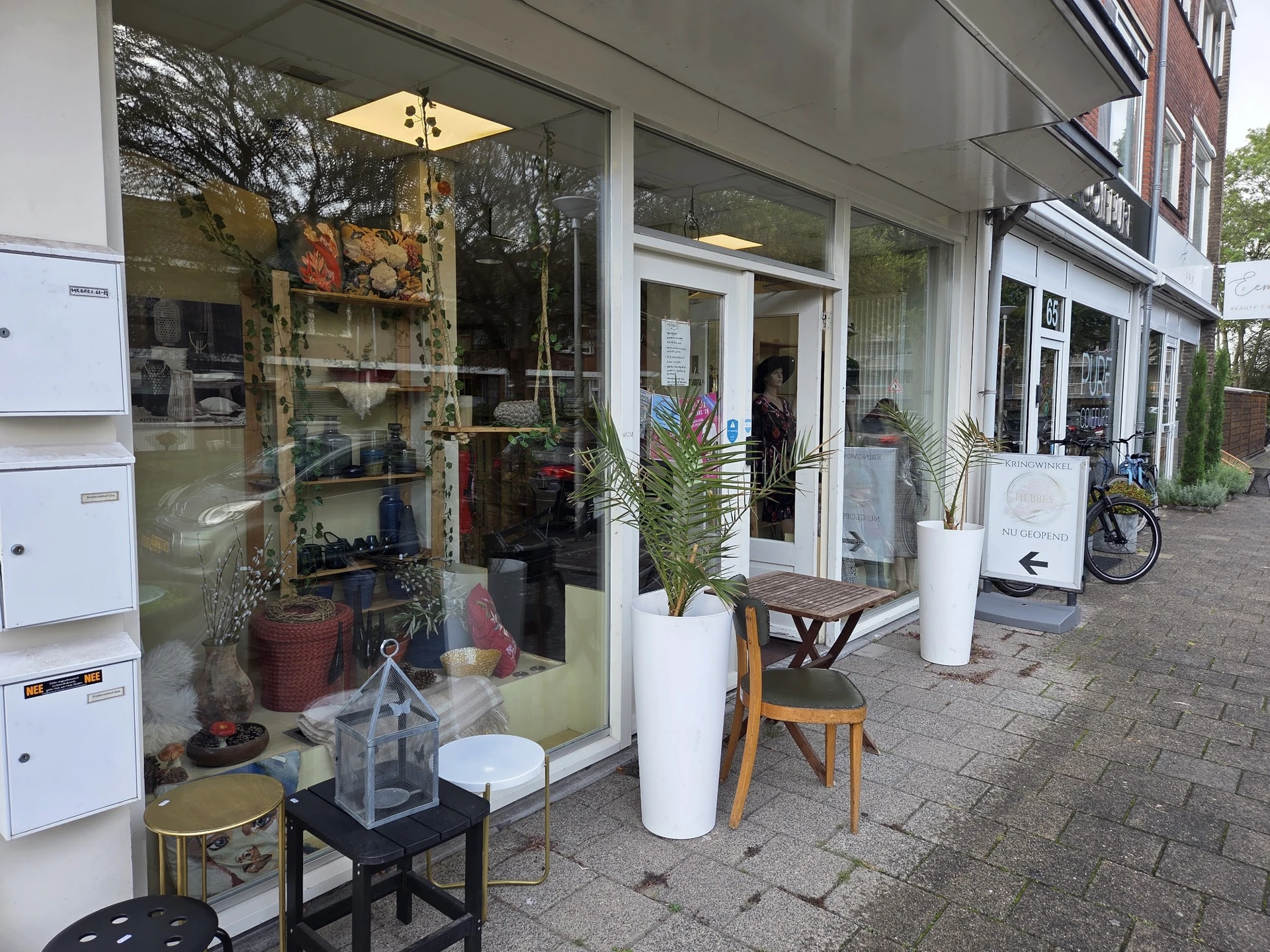 Winkelgevel met grote glazen ramen, witte deurkozijnen, plantenbakken en uitgestalde spullen op straat