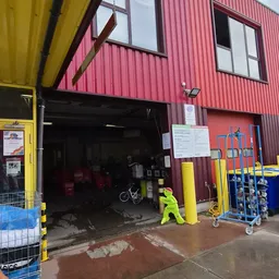 Ingang van kringloopwinkel met rode golfplaten gevel, gele details en diverse obstakels voor de deur