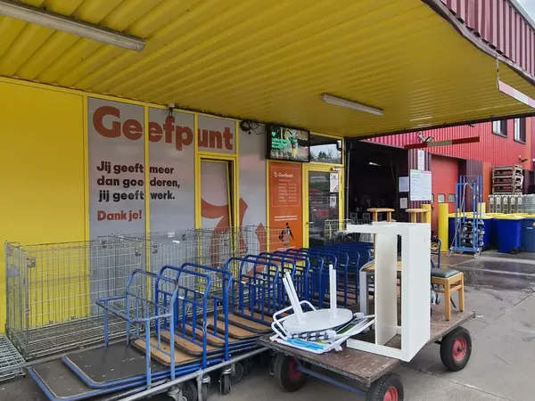 Gele gevel van kringloopwinkel met blauwe winkelwagens en rode deur, winkelname 'Geefpunt' zichtbaar