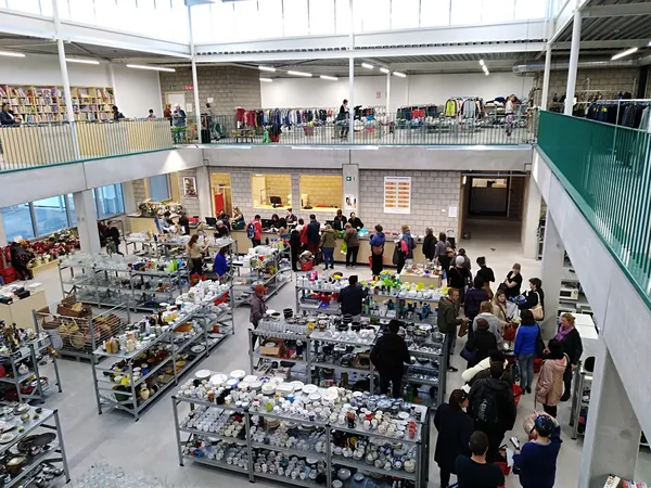 Overzicht van de kringloopwinkel met meerdere verkooptafels vol spullen, bezoekers op twee niveaus, stellingen met goederen t