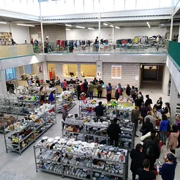Overzicht van de kringloopwinkel met meerdere verkooptafels vol spullen, bezoekers op twee niveaus, stellingen met goederen t