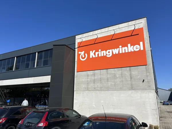 Voorgevel van kringloopwinkel met oranje logo en signage, parkeerplaats met auto's op voorgrond