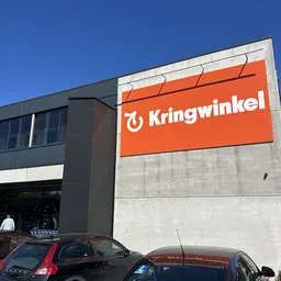 Voorgevel van kringloopwinkel met oranje logo en signage, parkeerplaats met auto's op voorgrond