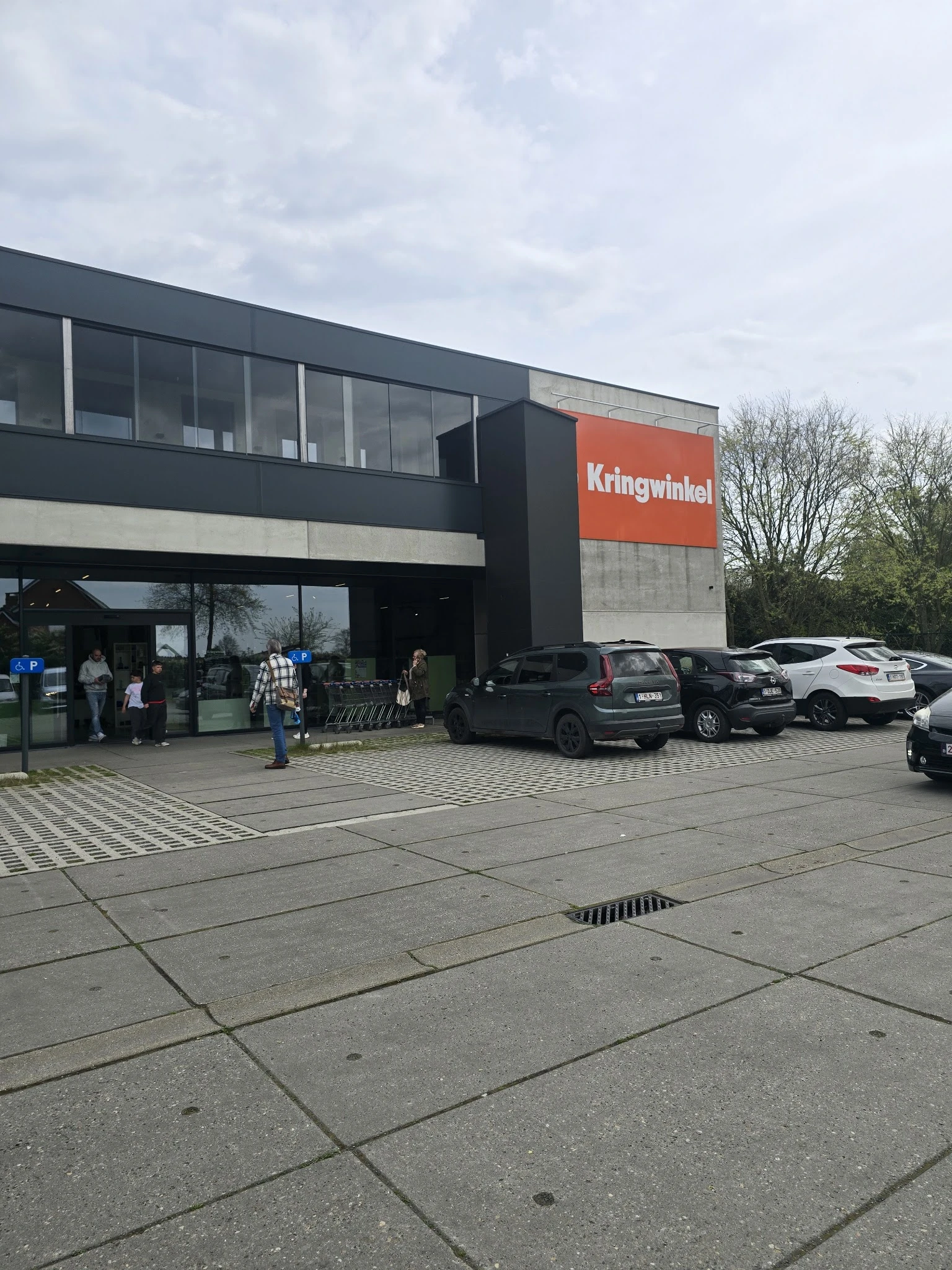 Moderne gevel van kringloopwinkel met oranje Kringwinkel-bord, parkeerplaats met auto's en klanten buiten