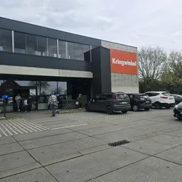Moderne gevel van kringloopwinkel met oranje Kringwinkel-bord, parkeerplaats met auto's en klanten buiten