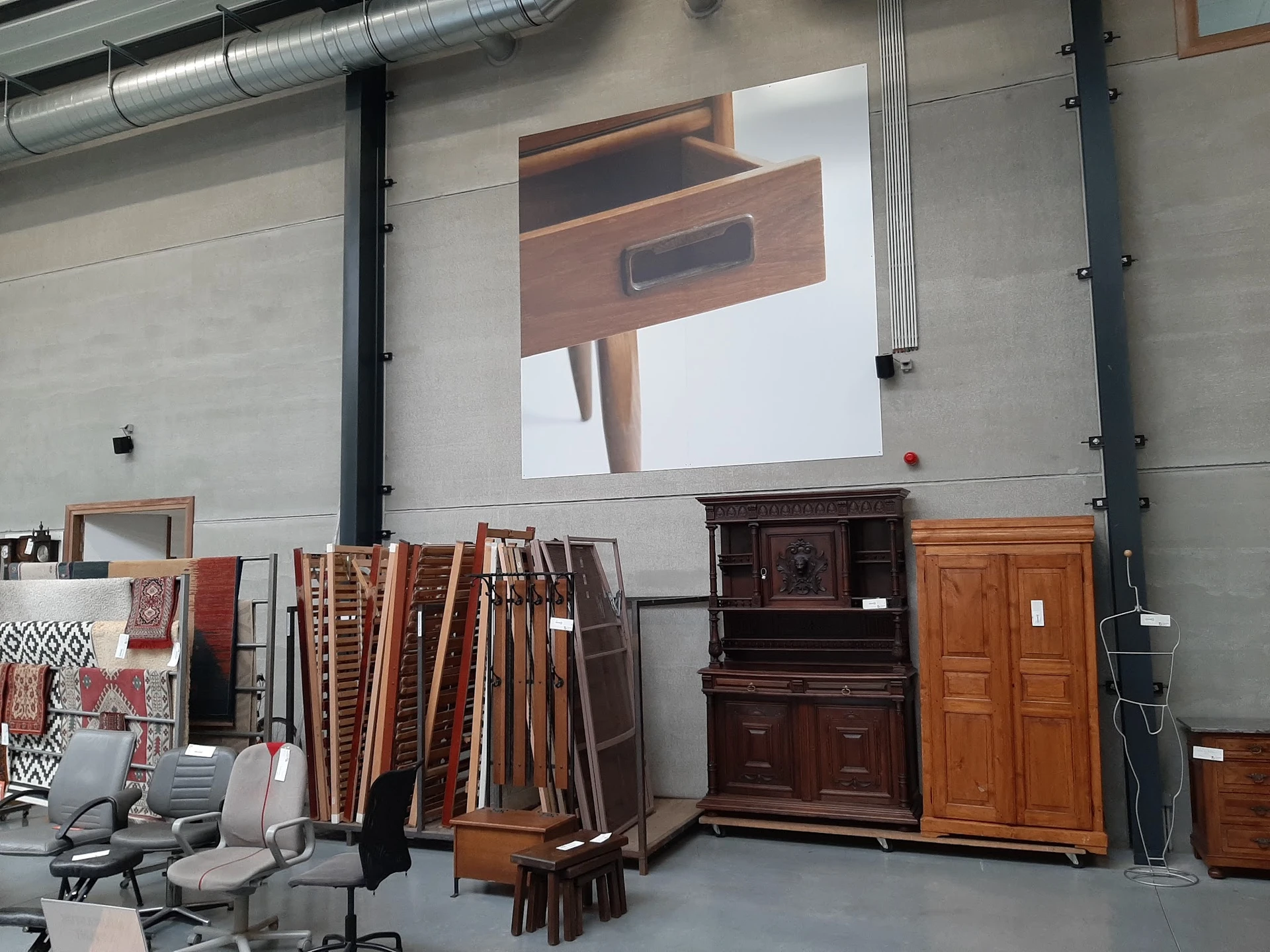 Meubels en huisraad in een kringloopwinkel, waaronder houten kasten, laden en stoelen, met groot projectiescherm aan de muur.