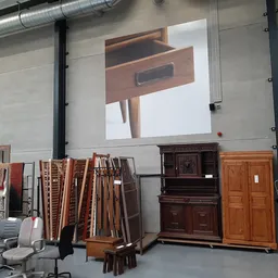 Meubels en huisraad in een kringloopwinkel, waaronder houten kasten, laden en stoelen, met groot projectiescherm aan de muur.