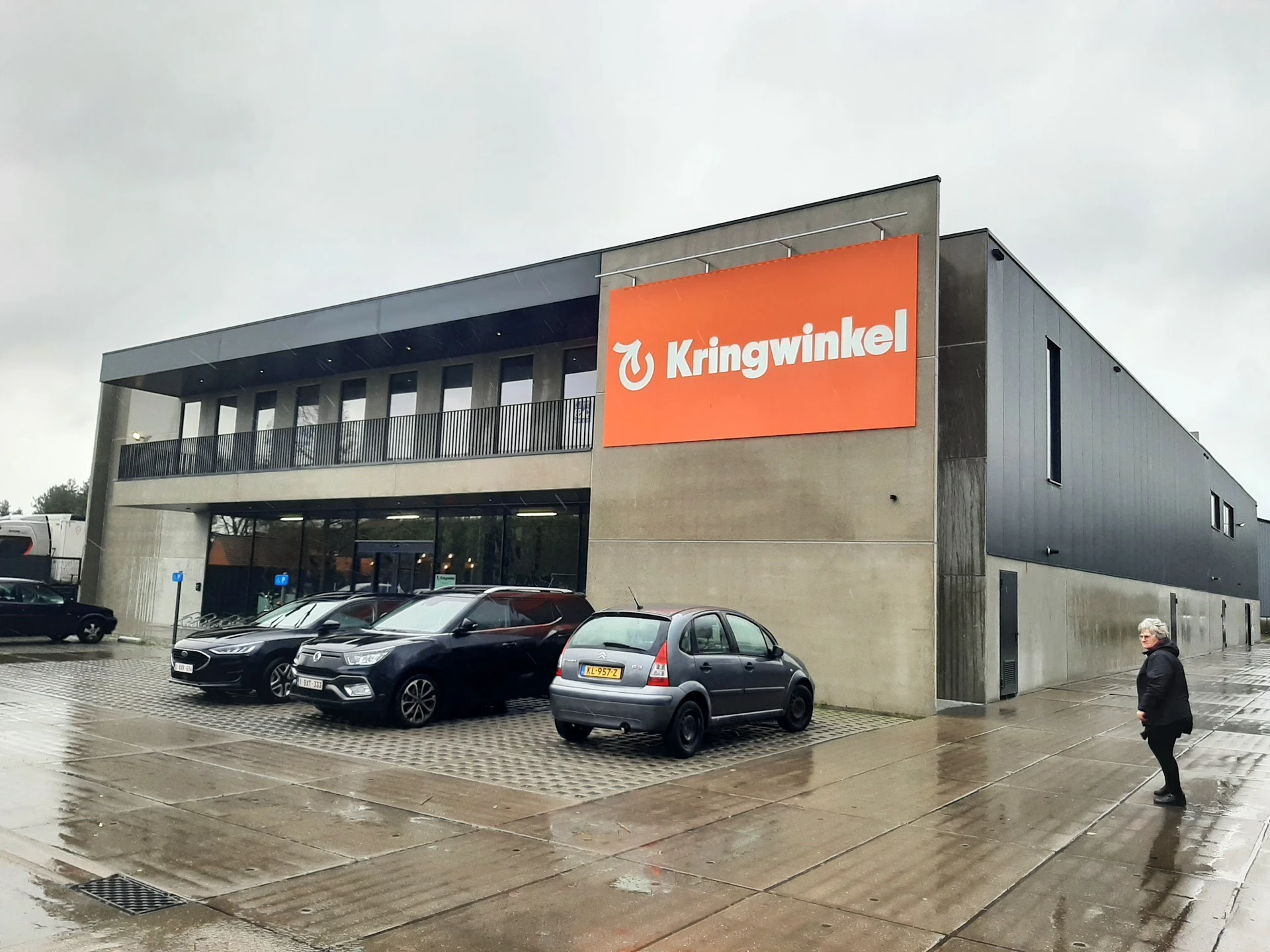 Moderne gevel van Kringwinkel met oranje logo, parkeerplaats met auto's, grijze bewolking