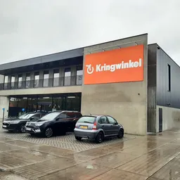 Moderne gevel van Kringwinkel met oranje logo, parkeerplaats met auto's, grijze bewolking