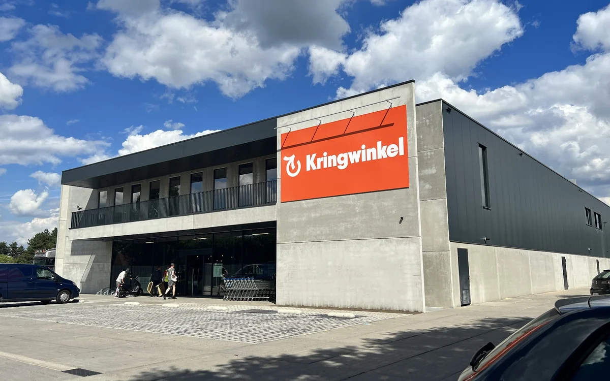 Modern grijs gebouw met grote oranje Kringwinkel-bord, parkeerplaats en blauwe lucht