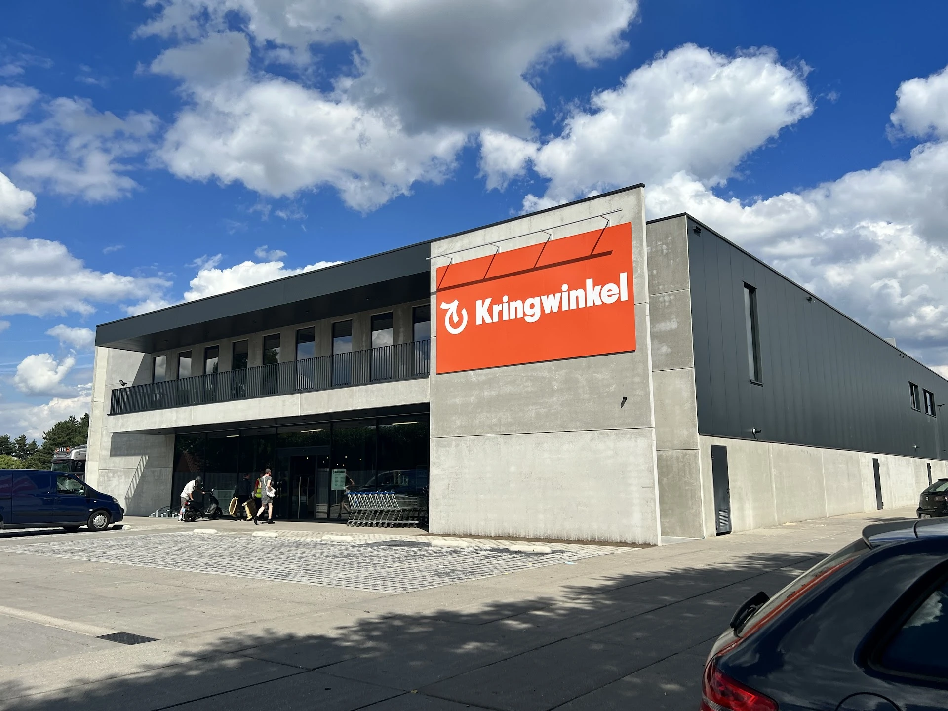 Modern grijs gebouw met grote oranje Kringwinkel-bord, parkeerplaats en blauwe lucht