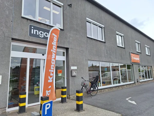 Grijze gevel van kringloopwinkel met witte ramen, oranje banner, parkeerplaats en fiets ervoor