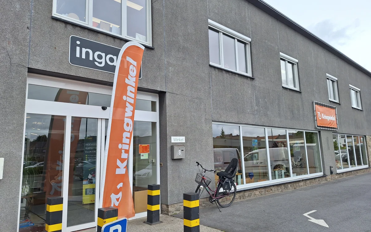 Grijze gevel van kringloopwinkel met witte ramen, oranje banner, parkeerplaats en fiets ervoor