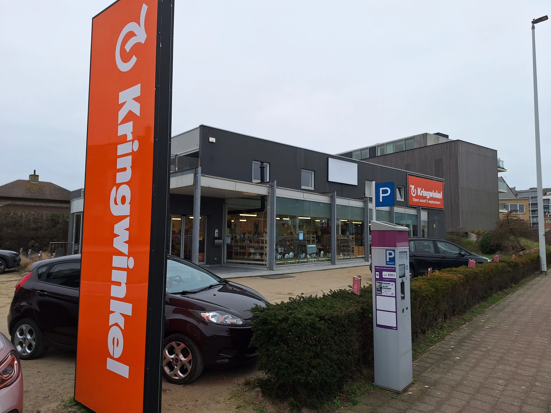 Voorgevel van kringloopwinkel met oranje logo, zwarte gevel, parkeerplaats en P-bord