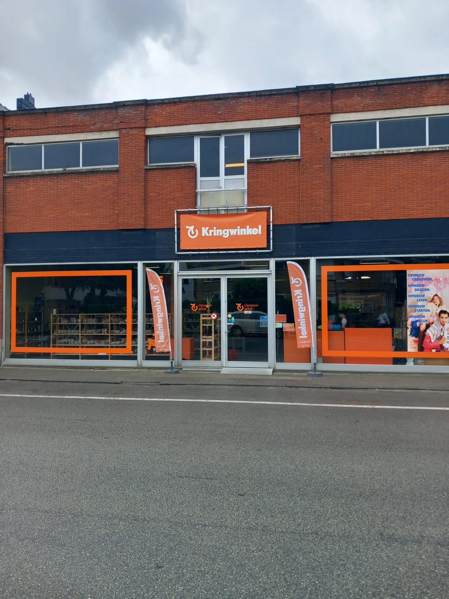 Gevel van kringloopwinkel met oranje en donkerblauwe accenten, etalages met spullen, duidelijk zichtbare winkelnaam