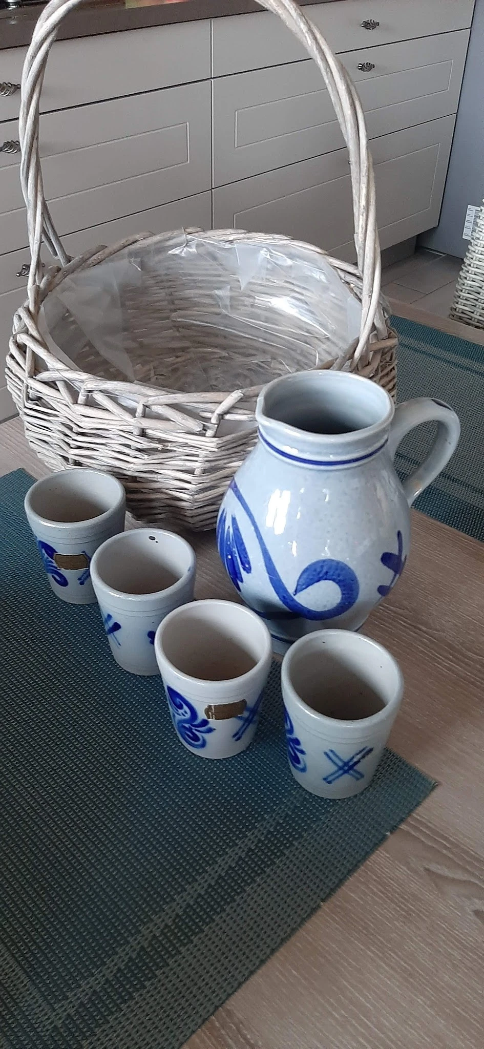 Blauwe en witte porseleinen kan met decoratief motief en vijf kleinere bekers, naast een rieten mand.