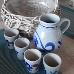 Blauwe en witte porseleinen kan met decoratief motief en vijf kleinere bekers, naast een rieten mand.