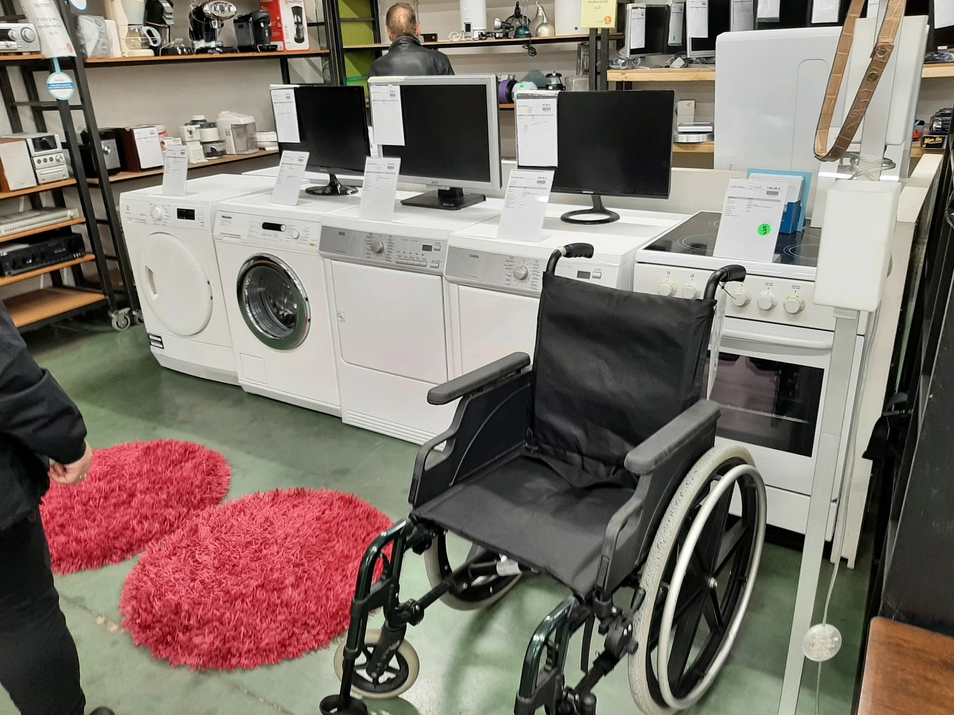 Winkelinterieur met witgoedapparaten, computermonitoren en een rolstoel op groene vloer