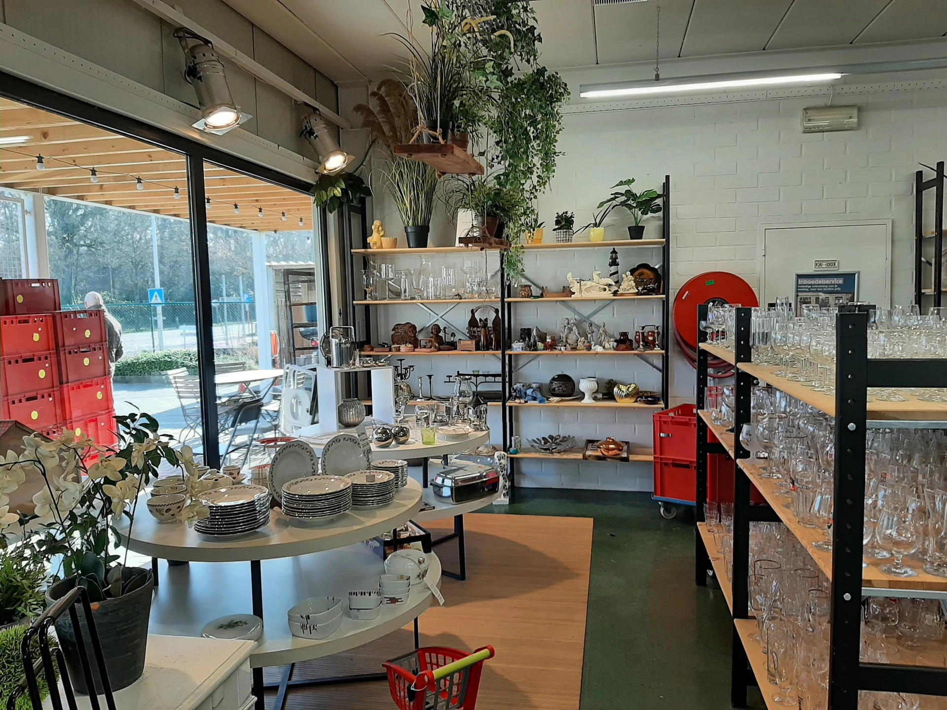 Winkelvloer met stellingen vol servies, glazen en decoratieve voorwerpen, ronde tafel met borden op voorgrond, planten en gro