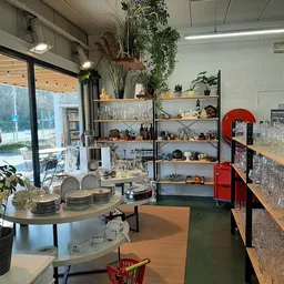 Winkelvloer met stellingen vol servies, glazen en decoratieve voorwerpen, ronde tafel met borden op voorgrond, planten en gro