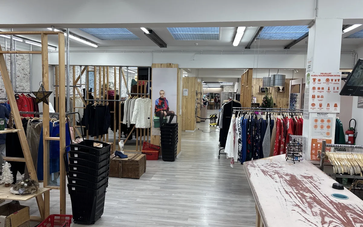 Ruim winkelinterieur met kledingrekken, houten stellingen en diversen tweedehandsartikelen verspreid over de vloer
