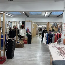 Ruim winkelinterieur met kledingrekken, houten stellingen en diversen tweedehandsartikelen verspreid over de vloer