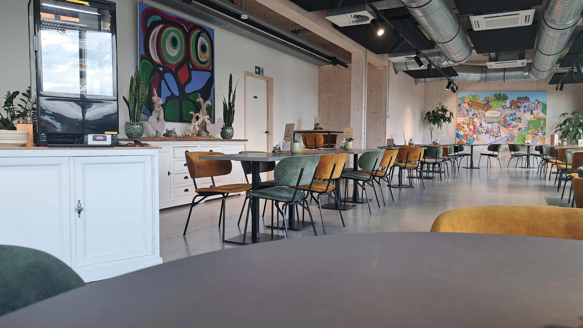 Ruime winkelruimte met houten stoelen aan tafels, kleurrijke kunstwerken aan de muur en planten als decoratie